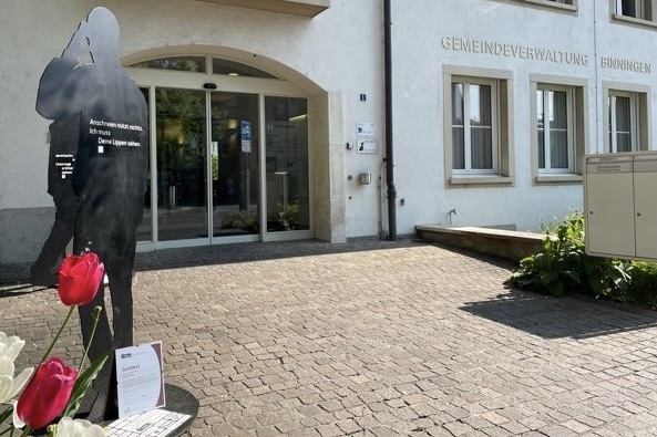Vor dem Eingang der Gemeindeverwaltung Binningen stehen zwei Silhouetten und das Zertifikat.