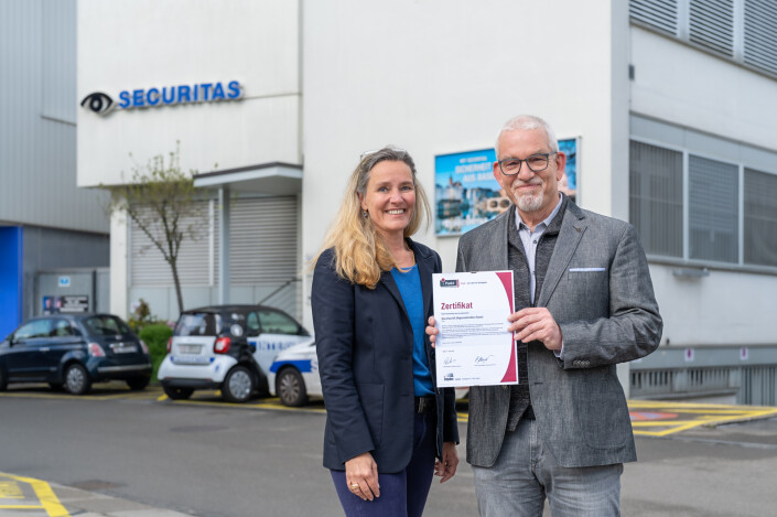 Daniela Reichenstein und Matthias Ph. Gnehm stehen nebeneinander und halten das iPunkt-Zertifikat in die Kamera. Im Hintergrund ist das Gebäude der Securitas AG, Regionaldirektion Basel zu sehen.